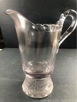 Front view of the pressed glass pitcher showing clear to light purple tint, scalloped rim edge, and lion motif on the base.