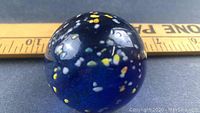 Close-up photo of large cobalt blue marble with embedded yellow and white mica flakes, measuring approximately 2.25 inches in diameter shown with ruler