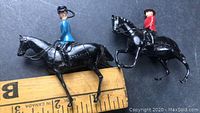 Two lead toy soldiers on black horses, one in blue the other in red, placed next to a ruler for size reference.