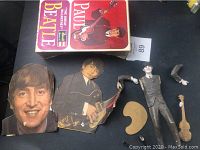 Photo showing Beatles paper cutouts of John Lennon and George Harrison, plastic arms, small guitar model piece, and one-sided Paul McCartney Beatles box.