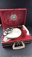 Full view of the vintage portable phonograph record player with red, white, and blue striped case, open lid showing the red interior with white artistic illustrations and a picture of cranes.