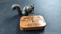 Photo showing the cold carved soapstone squirrel figurine beside the vintage mustard yellow metal tin for Mustad-Kirby fish hooks.