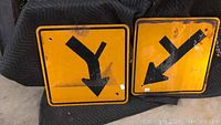Two yellow road signs with black directional arrows laying on a protective blanket showing general wear and rust spots.