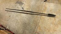 Two long forged spears laying on the floor, showing overall length and shape with pointed spearheads at one end and handles at the other.