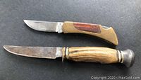 Both folding knives displayed open, showing blades and handles, one brass and wood, the other antler with metal bolster.