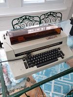 Front view of the cream-colored IBM Selectric II typewriter with an orange-brown translucent cover over the typing area, showing keyboard and overall condition.