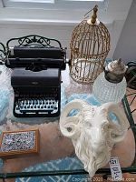 Overview of all items on glass table by window: vintage Royal typewriter, decorative birdcage, pressed glass ewer, porcelain ram head, wooden box with metal lid