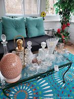 Photo of glassware and decanters on a table with cushions in the background, featuring four clear glass decanters with stoppers, gold cocktail shaker, six shot glasses, and a large egg-shaped Ethan Allen box.
