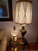 View showing both lamps on a glass-topped side table beside a sofa, with the tall brass lamp having a cream silk shade and the smaller lamp lit illuminating the glass crystal ball and bead details.