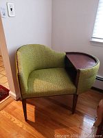 Full side angle showing the green fabric upholstery, metal stud trim, wooden legs, and curved shape of the chair.
