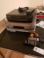 Stacked electronics including Quasar VHO 940 VHS player on top, Sonic Blue VHS/DVD combo below, and Sony DVR at the bottom with associated remotes and headphones on top