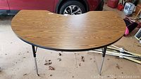 Kidney-shaped table with faux wood grain laminate surface, black edging, and four metal legs. The two front legs are silver with adjustable height features.