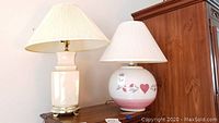 Two ceramic base lamps on wooden surface against white wall. Left lamp is beige rectangular with brass pedestal and pleated shade. Right lamp is spherical with painted floral and heart design and white pleated shade.