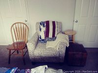 View of room corner showing easy chair with script print upholstery with three throws draped on it, a wooden spindle back chair, a round wooden stool, and leather ottoman on carpeted floor.