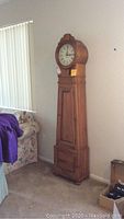 Full front view of the wooden grandfather clock standing next to a sofa.