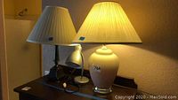 Three lamps on wooden surface: ceramic beige urn-shaped lamp with pleated shade, dark metal lamp with pleated shade, small silver desk lamp