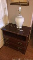 Wood end table with two drawers and open shelf, showing gold-tone handles and dark brown finish in a corner next to the wall.