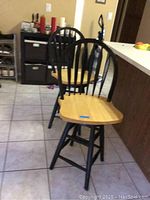 Two wooden bar stools shown side by side on tiled floor with natural wood seats and black spindle backs and legs.