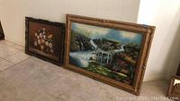 Two framed canvas prints leaning against a wall on a carpeted floor, one large nature scene with waterfall and gazebo, the other floral arrangement still life.