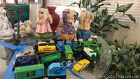 Photo shows three ceramic bunny figurines and multiple toy train cars and die cast cars on a glass surface surrounded by houseplants and vases.