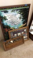 Three framed pieces leaned against wall: large Inspirational Psalm print with nature background and text, framed Last Supper print, and wooden carved Last Supper plaque.
