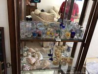Wide view of glass shelves showing collection of shot glasses, cocktail glasses, and sea shells inside wooden curio cabinet with mirror back
