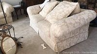 Whole view of the beige cloth upholstered love seat with floral damask fabric and two cushions next to a mirror, showing curved armrests and skirted base.