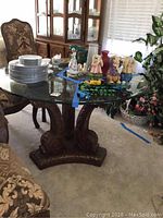 Full view of the square glass top pedestal dining table with stacked plates on top for size reference. Shows the detailed carved wooden base and surrounding upholstered chairs.