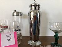 Photo showing four branded vintage beer glasses and a polished stainless steel cocktail shaker with lid on a wooden surface.
