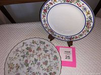 Two decorative china plates displayed on a quilted surface, one with blue rim and floral border, the other with scalloped edge and delicate floral pattern.