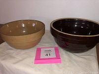 Two nesting ceramic bowls, one glazed dark brown with cream rim, the other unglazed tan. Both marked with numbers on the bottom.