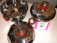 Two vintage aluminum serving dishes with Bakelite handles and lids, showing the reflective aluminum and bright red details