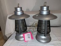 Two silver plug-in bug zappers placed side by side on a covered surface, showing their full height including the flat top and black grid, with power cords trailing behind.