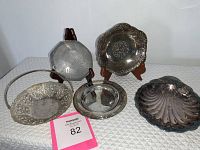 Lot of five silver items on white textured tablecloth, including a bride's basket, silver bowls, and a shell dish, some on wood stands.