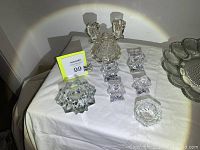 Various pressed glass items including candlesticks, votive holders, deviled egg server, and candy dish on white cloth.