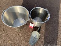 Photo of two stainless steel milk pails side by side with metal grain scoop on pavement, showing overall size and condition.