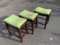 Three modern barstools arranged outdoors against gray pavement background showing green leather seats with nailhead trim and dark wooden squared legs