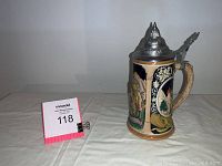 Side view of vintage German beer stein showing painted ceramic body and pewter lid with thumb lever on a white cloth background.