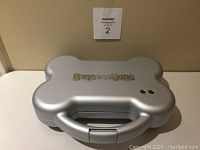 Closed view of silver dog treat maker shaped like a bone with logo on top and two indicator lights.