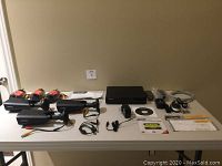 Photo shows three black metal Defender security cameras with cables, a black rectangular Defender DVR unit, a remote control, a computer mouse, power adapters, a software CD and instruction manuals all laid out on a table.