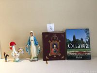 Lot photo showing all items: 8" Portuguese rooster, 4" metal rooster bottle cork, 12" Virgin Mary ceramic statue, Holy Bible, and Ottawa book.