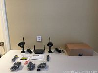 Wide shot of all items displayed on white table including two wireless cameras, receiver, cables, power adapters, quick guide, warning stickers, and box.