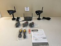 Full set of Defender Phoenix Pro wireless security camera system components displayed on table: two cameras with stands and antennas, wireless receiver, power adapters, connection cables in plastic, user manual, and box.