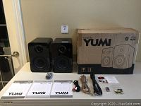 Two black gloss Kanto YUMI speakers with remote, manuals, cables and box on table