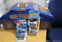 Photo showing the Hot Wheels soft fabric case shaped like a blue car with Hot Wheels logo and flames, along with 6 new Hot Wheels cars in original sealed packages in front of the case.