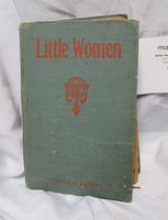 Front cover of the antique Little Women book showing the green cloth cover with red text and symbol, worn edges, and visible damage to the binding and pages.