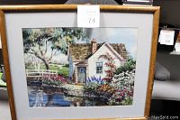 Framed print depicting a cottage by the water with garden and trees in full view.