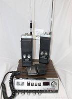 Two Realistic TRC-220 walkie talkies with antennas upright behind Sears RoadTalker 40 base station and handheld microphone on wood grain case.