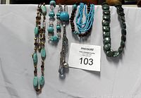 Arrangement of 5 assorted stone bead necklaces on white cloth background showing different bead shapes and colors including turquoise, brown, and greenish blue.