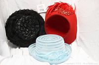 Three hats displayed: black woven hat with netting, red felt hat with feather and applique details, light blue transparent rimmed hat with floral decoration